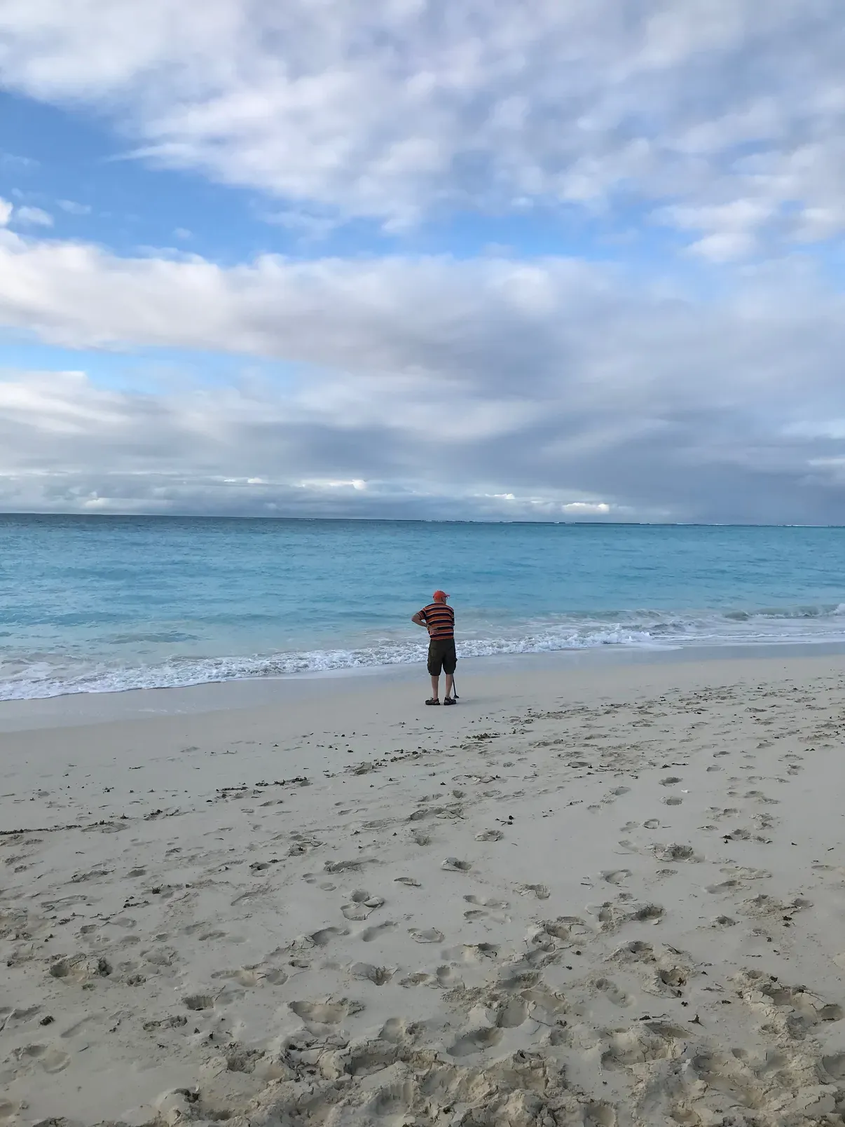 A single figure standing at the waterline of a vast empty beach, turquoise sea beyond