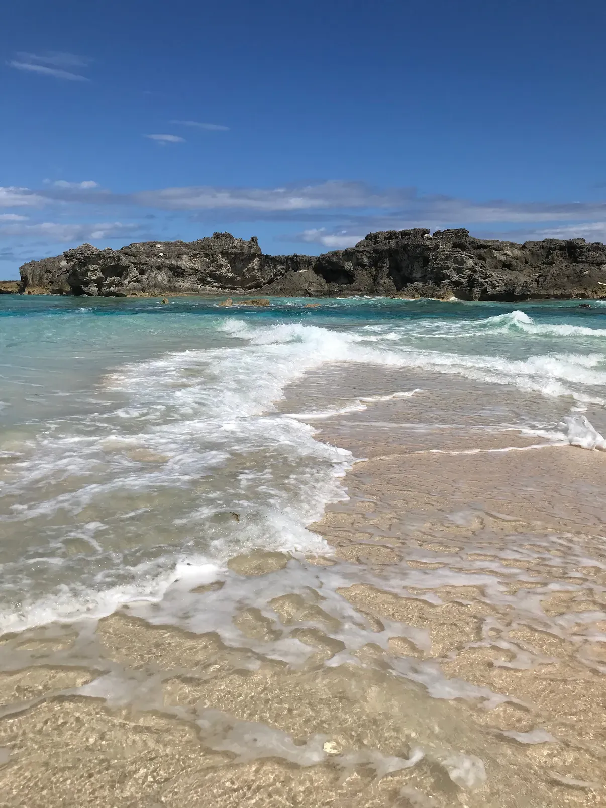 Foamy waves washing over pale sand with a rocky headland behind, North Caicos