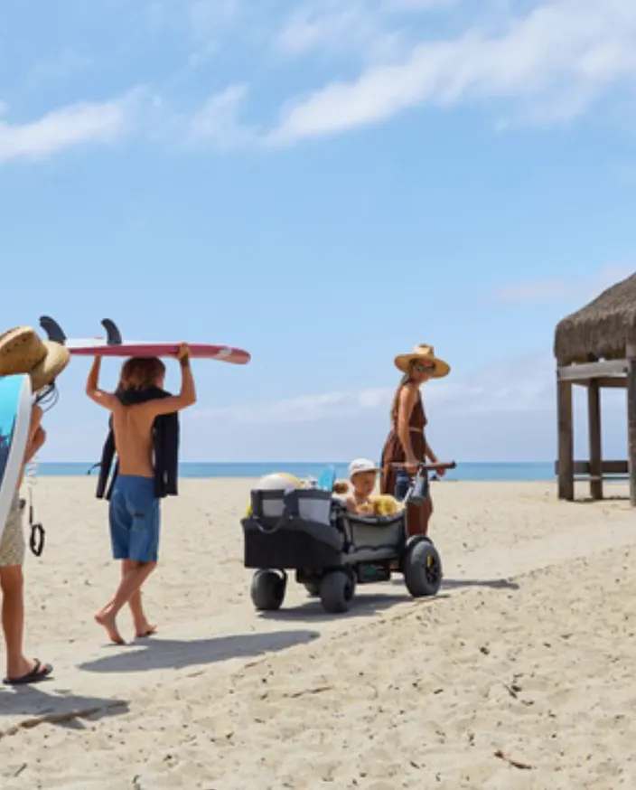 A Veer Cruiser on beach wheels crossing soft sand with a kid riding inside, surfers walking past. Photo from Veer.